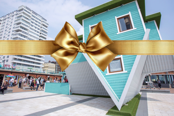 Upside-down turquoise house with gold ribbon and bow; people and buildings in background.