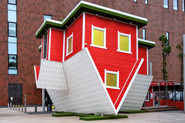 Upside-down red house with white trim on urban street with modern building backdrop.