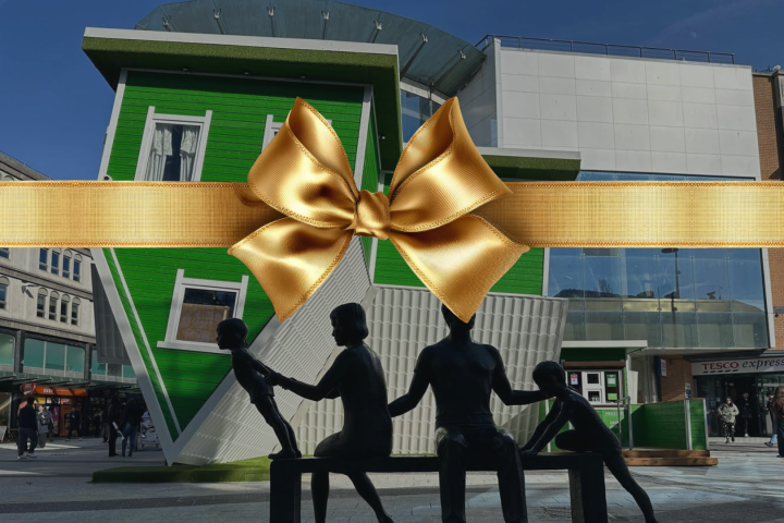 Upside-down green house with gold ribbon and seated figures statue in front.