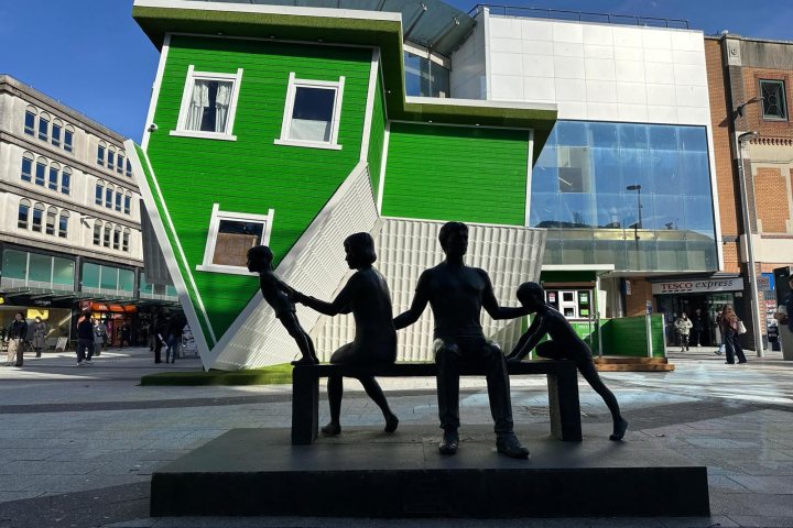 Sculpture of people on a bench in front of an upside-down green house in a plaza.