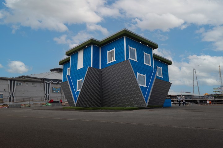 Upside-down blue house with a green roof, under a cloudy sky.