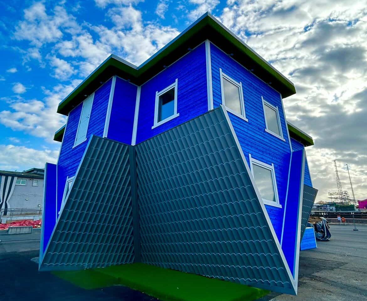 Upside-down blue house with green roof, set against a cloudy sky.